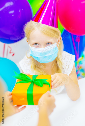 A girl in a festive hat and a medical mask on her face is congratulated on her birthday by holding out a wrapped gift. Birthday during a coronavirus or disease pandemic
