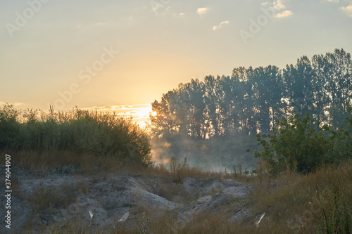 Fototapeta Naklejka Na Ścianę i Meble -  Summer time. Morning dawn over the river in a hazy, thoughtful haze. Beautiful view of the forest and river covered with fog early in the morning.