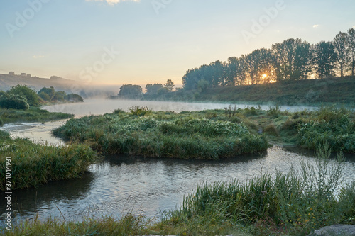 Summer time. Morning dawn over the river in a hazy, thoughtful haze. Beautiful view of the forest and river covered with fog early in the morning.