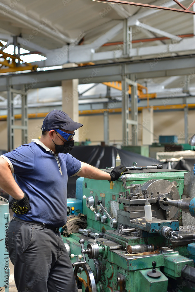 Working turner wearing a protective antiviral mask on a lathe in a workshop. The machine operator at the workplace controls the equipment.