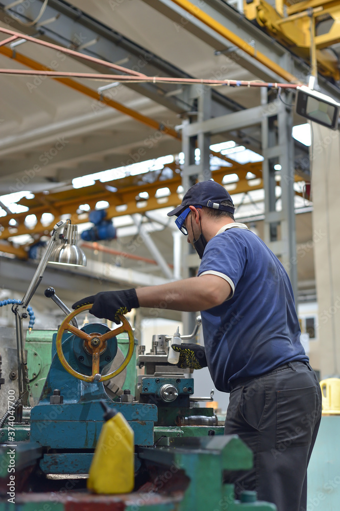 Working turner wearing a protective antiviral mask on a lathe in a workshop. The machine operator at the workplace controls the equipment.