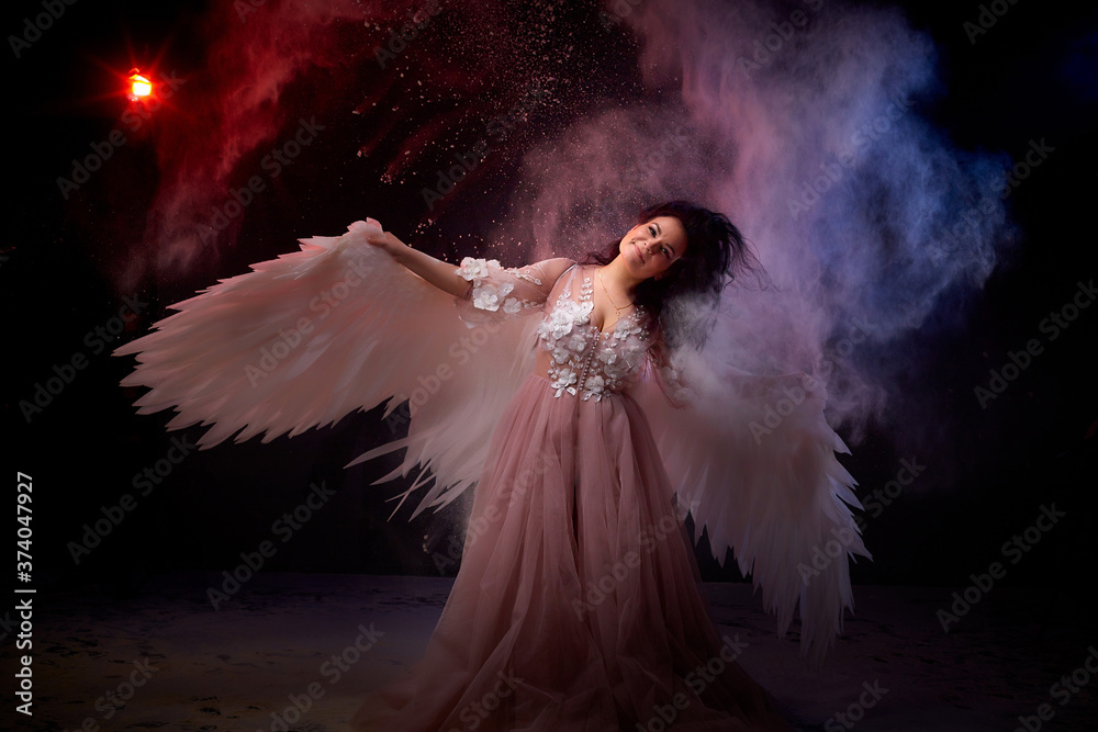 Naklejka premium Girl with white wings posing in dark black studo during photoshoot with flour or dust and light. White angel in dark space