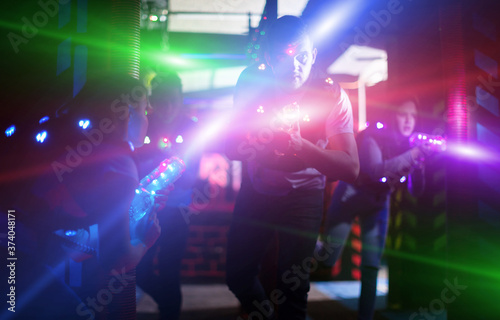 Emotional guy playing laser tag with friends on dark labyrinth in colorful beams of laser pistols