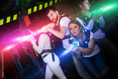 Modern young people playing laser tag on dark labyrinth in bright beams of laser pistols