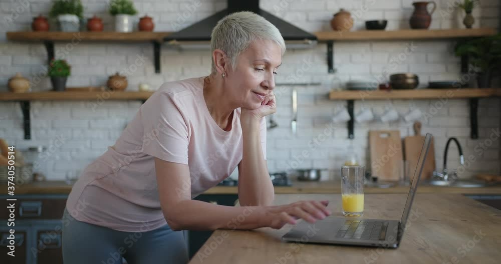 Portrait of elderly woman in pink t-shirt standing at the kitchen and using laptop Portrait of elderly woman in pink t-shirt standing at the kitchen and using laptop