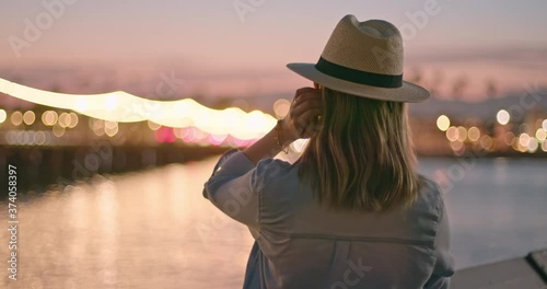 Slow motion of woman enjoying view on promenade lighting. Back view of stylish young female traveler in hat enjoying evening on the pier with night city view. Female traveler exploring USA coast
