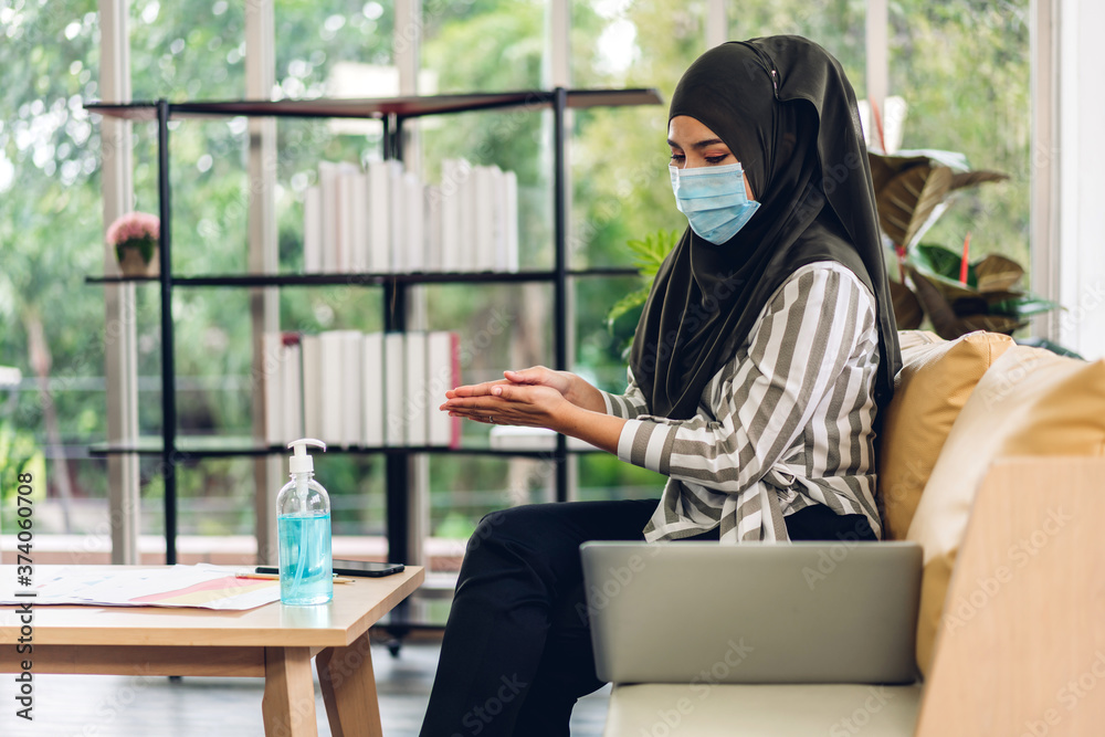 Asian muslim woman washing hands with alcohol gel in quarantine for coronavirus wearing protective mask with social distancing and using laptop computer working at home.work from home and covid19 Asian muslim woman washing hands with alcohol gel in quarantine for coronavirus wearing protective mask with social distancing and using laptop computer working at home.work from home and covid19
