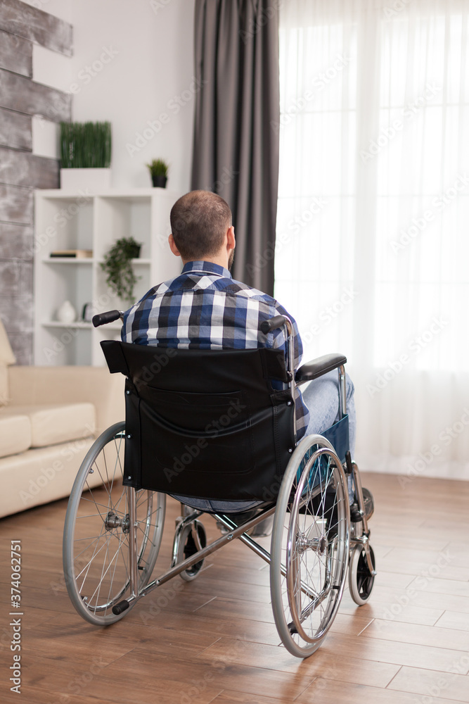 Back view of man with walking disability sitting on wheelchair. Serious sad caucasian man wearing casual clothes and look at large panoramic view in bright modern living room, hospital or clinic. Back view of man with walking disability sitting on wheelchair. Serious sad caucasian man wearing casual clothes and look at large panoramic view in bright modern living room, hospital or clinic.