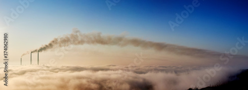 Panoramic view of tops of three smoking stacks of thermal power station on the horizon taken from the hill, pipes in morning fog on blue sky, concept of energy generation, ecology and pollution