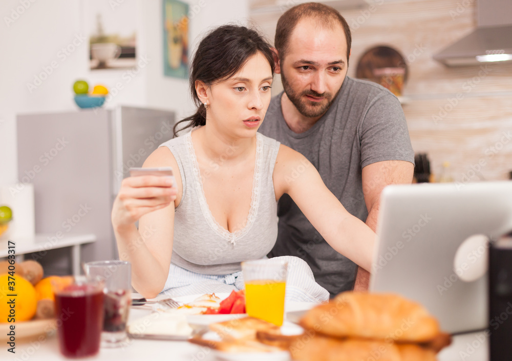 Young couple using laptop at the kitchen. Happy married husband and wife in pajamas using internet web online modern technology, smiling and happy in the morning. Reading news browsing internet