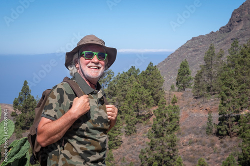 Smiling senior man with hat and backpack enjoying excursion in mountain range in Tenerife island - concept of summer vacation for an elderly active people