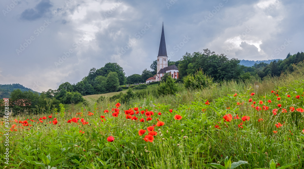 Alpine landscape. Scenic view of catholic church on the hill, meadow with wild poppies in the foreground. Travel Slovenia. Alpine landscape. Scenic view of catholic church on the hill, meadow with wild poppies in the foreground. Travel Slovenia.