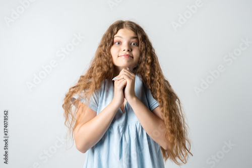 Young woman with curly long hair keeps hands together near chin, smiles gently, has cute expression, wears blue t-shirt
