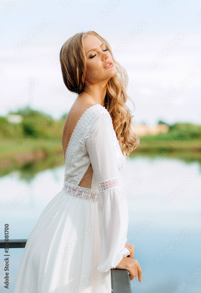 Blonde hair bride in white silk long lace dress standing on the terrace with lake view