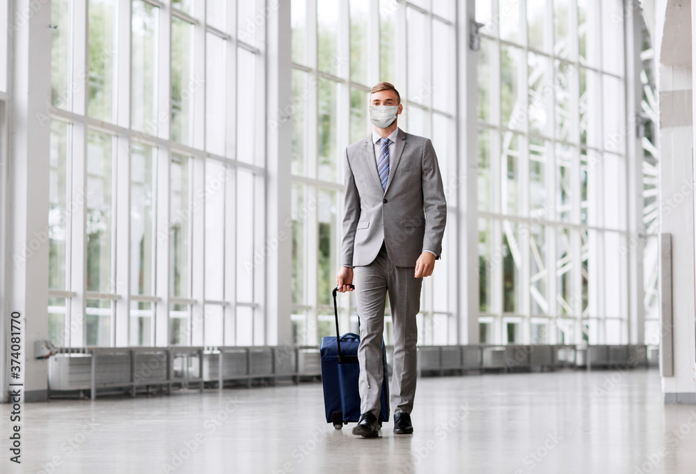 business trip, health and people concept - young businessman walking with travel bag along office building or airport wearing face protective medical mask for protection from virus disease