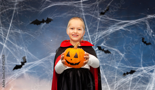 halloween, holiday and childhood concept - happy smiling girl in black dracula cape or costume with jack-o-lantern pumpkin over bats and cobweb in night sky on background © Syda Productions