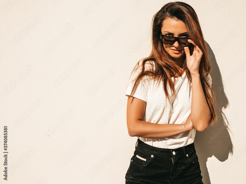 Portrait of beautiful model in sunglasses. Female dressed in summer hipster white T-shirt and jeans. Trendy woman posing near wall in the street Portrait of beautiful model in sunglasses. Female dressed in summer hipster white T-shirt and jeans. Trendy woman posing near wall in the street