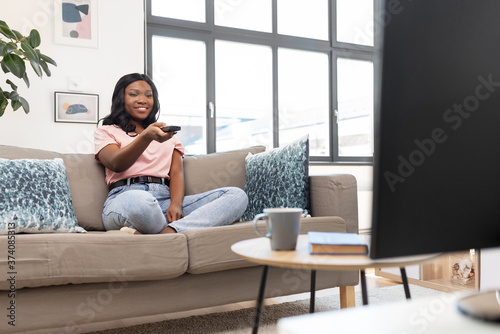 people and leisure concept - happy smiling african american young woman with remote control watching tv at home