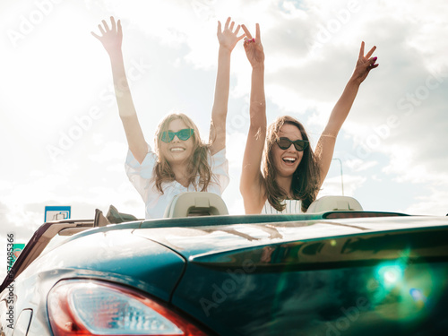 Portrait of two young beautiful and smiling hipster women in convertible car. Sexy carefree female driving cabriolet. Positive models riding and having fun in sunglasses. They raising hands