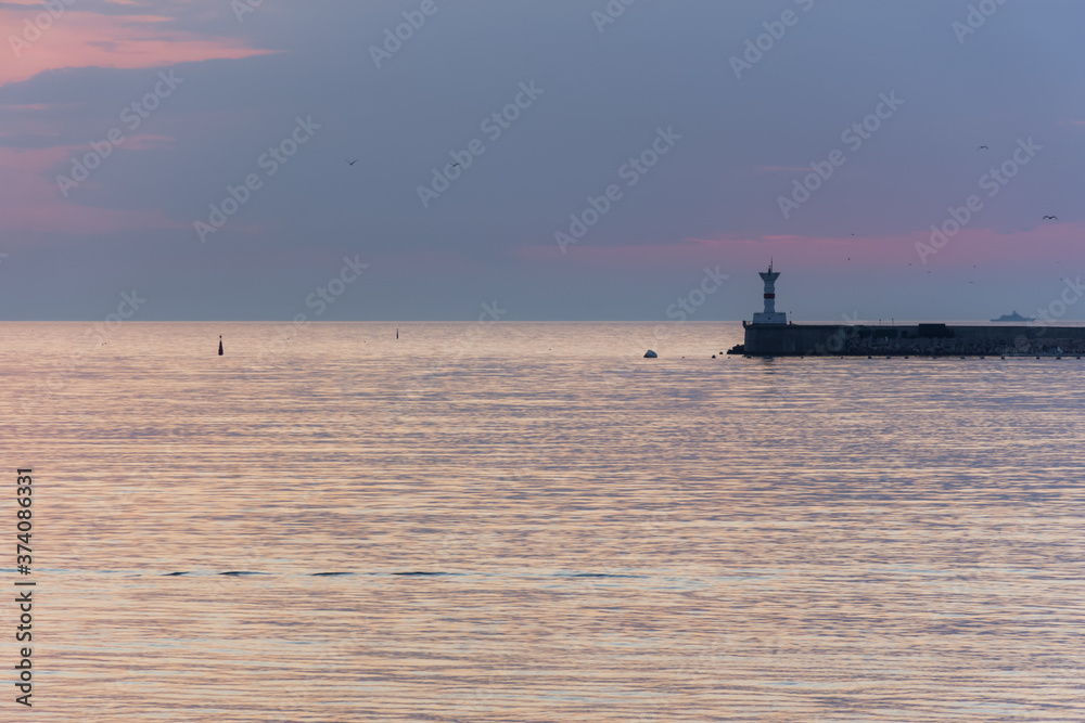 A lighthouse against the sunset sky. Beautiful peaceful seascape. Small ships on the sea horizon. Purple clouds and soft pink water. Seaport. Gulls in the sunset sky. Atmospheric evening landscape.