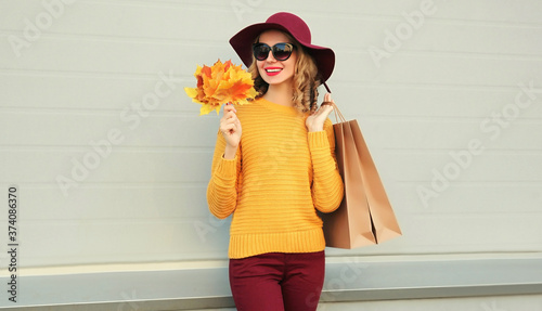 Autumn portrait of woman with shopping bags and yellow maple leaves wearing a sweater, hat over gray background