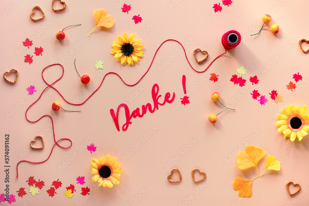 Obraz premium Danke means Thank you in German language. Fall decorations - yellow flowers, orange gingko leaves, maple leaf paper confetti, heart shape pretzel snacks. Autumn flat lay on orange background.