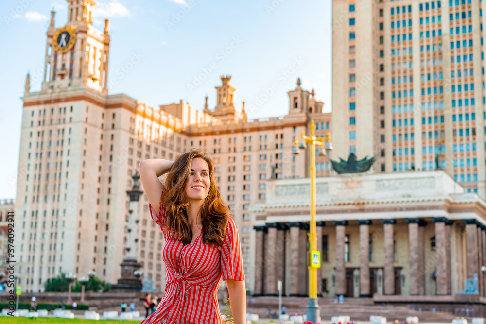 Obraz premium Cute young woman in a red dress poses against the background of the main University of Russia, Moscow high-rise