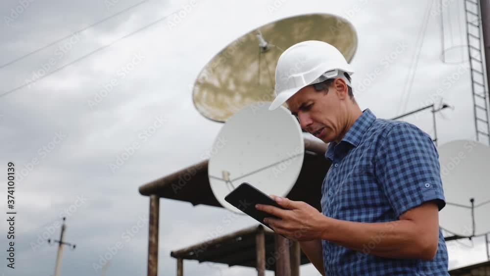 Helmet engineer and with a tablet standing on the roof of the building working with a satellite dish. Successful quality performance of tasks. Helmet engineer and with a tablet standing on the roof of the building working with a satellite dish. Successful quality performance of tasks.