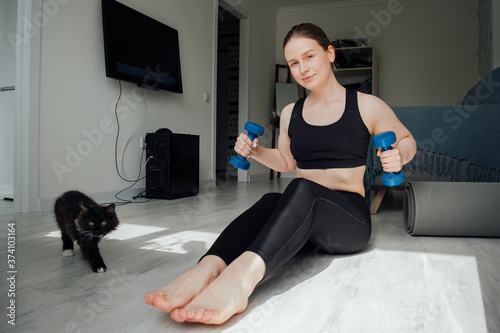 the girl is engaged with dumbbells sitting on the floor at home. doing sports at room. the cat walks by the owner. apartment interior background. Positive life