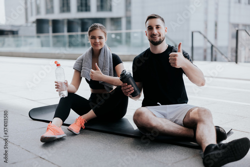 happy young couple brunette beautiful woman and handsome man in sportswear show like sign, stretching on yoga mat, drinking water , healthy fit lifestyle, street workout photo