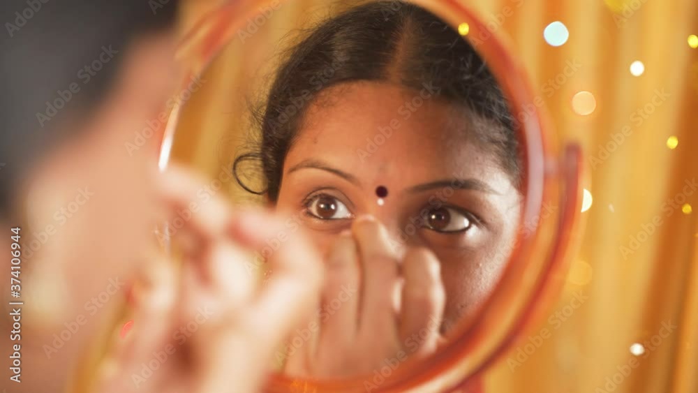 Indian married Woman applying Bindi, sindoor or decorative mark to forehead in front of mirror during festival celebrations with decoration lights as background