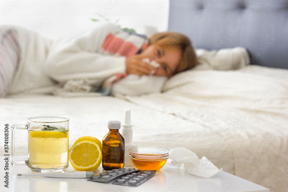 Selective focus on alternative medicine on the night table (hot drink, lemon, honey, pills, thermometer, nasal spray and crumpled handkerchiefs. Young sick woman laying on bed Selective focus on alternative medicine on the night table (hot drink, lemon, honey, pills, thermometer, nasal spray and crumpled handkerchiefs. Young sick woman laying on bed
