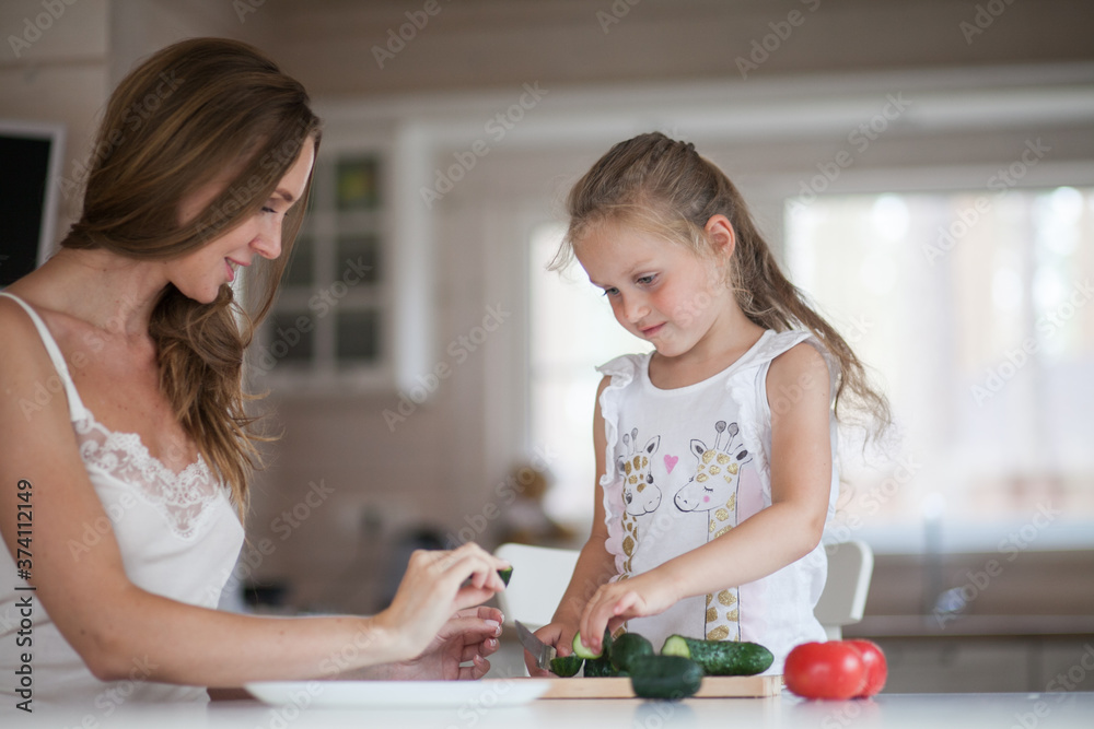 Naklejka premium Happy family mother and child posing at home. Beautiful young mom and little daughter having fun and preparing vegetables for salad in a white kitchen in a Scandinavian style interior. Healthy food.