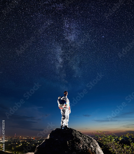 Space traveler standing on top of rocky hill and looking at beautiful sky with stars and Milky way. Astronaut in white space suit holding helmet. Concept of space travel and cosmonautics.