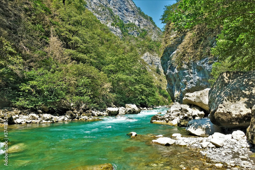 The mountain river flows through the gorge between the rocks. The turquoise water is foaming. There are boulders on the banks. Green vegetation on the steep slopes. Summer sunny day.