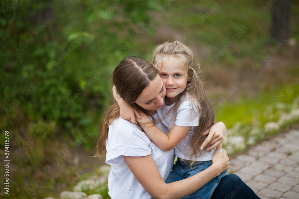 Happy smiling mother and her cute emotional little daughter child in white shirts and denim jeans are hugging and having fun outdoor in nature at countryside. Spending summer time with family. Happy smiling mother and her cute emotional little daughter child in white shirts and denim jeans are hugging and having fun outdoor in nature at countryside. Spending summer time with family.
