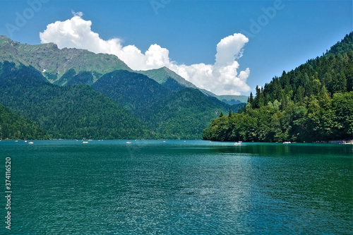 Alpine lake Ritsa. Boats float on the smooth turquoise water. The mountain slopes are covered with dense forest. There are picturesque clouds in the azure sky. Abkhazia.