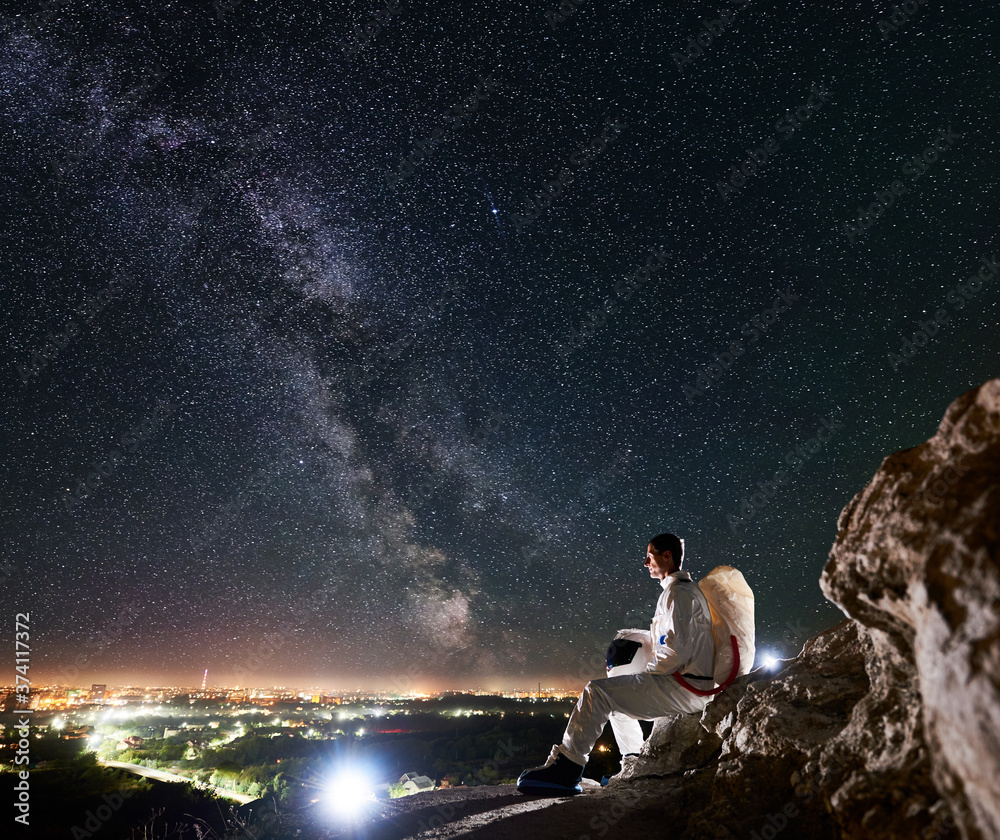 Obraz premium Spaceman traveler sitting on rocky mountain under fantastic starry sky. Space traveler enjoying beautiful view of Milky way and night city in the valley. Concept of cosmonautics and space travel.