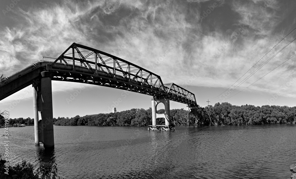 Beautiful black and white panoramic view of a river with reflections of a tall pedestrian and water pipe bridge, trees and sky on water, Parramatta river, Rydalmere, Sydney, New South Wales, Australia