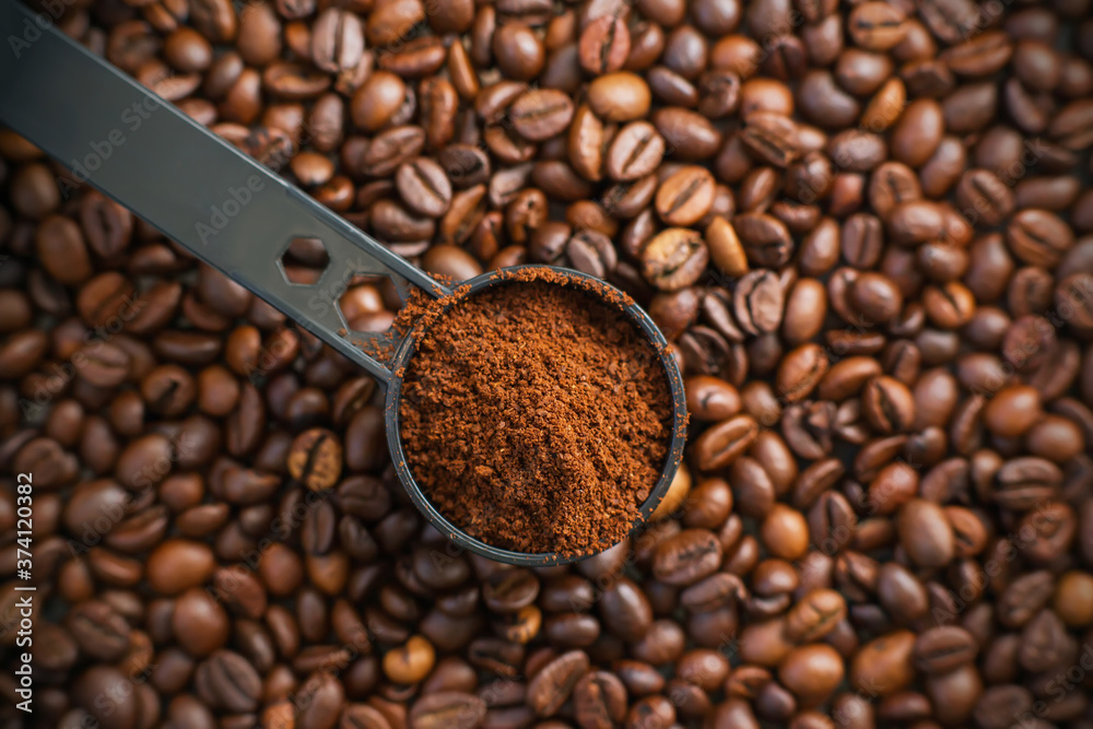 Fototapeta premium Against the background of roasted aromatic coffee beans lies a long black measuring spoon, which is filled with ground coffee. Detail from the coffee maker. The process of making coffee.