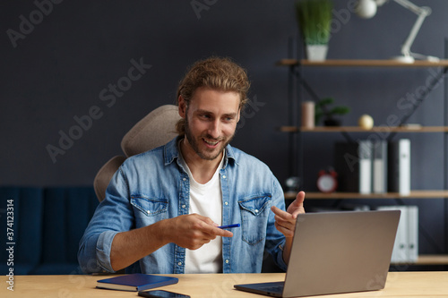 Studying online,online courses.Business concept.Young handsome curly smiling man with long hair studying in home with laptop.Business portrait of handsome manager sitting at workplace.