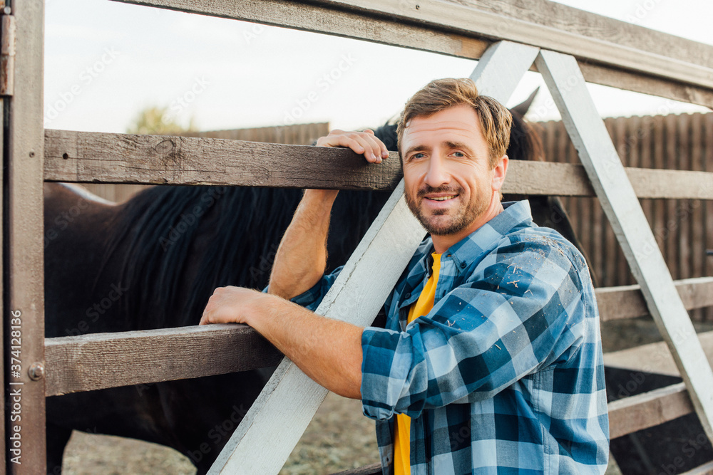 farmer in checkered shirt looking at camera while leaning on corral fence near horse farmer in checkered shirt looking at camera while leaning on corral fence near horse