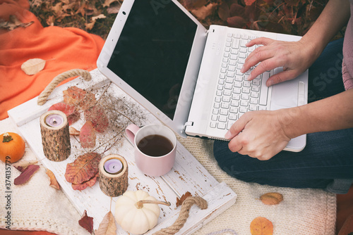 The girl works at a laptop on a red blanket in the autumn garden. Next to it is a tray with a cup of tea and autumn leaves and pumpkins.