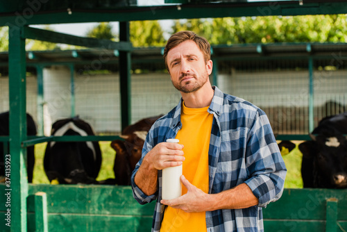 rancher in checkered shirt holding bottle of fresh milk while looking at camera near cowshed