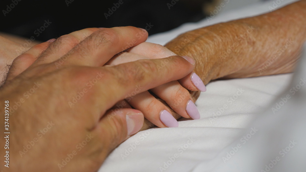 Fototapeta premium Granddaughter and grandson gently touch hand of their grandma lying in bed hospital. Young woman and man comfort wrinkled arm of sick grandmother in medical clinic. Concept of care and love. Close up