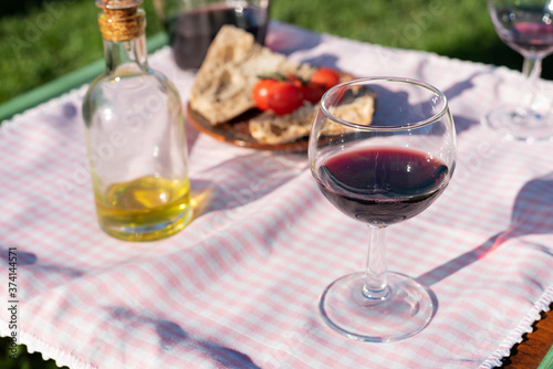 Red wine cup and olive oil picnic in garden at sunset. Blurred background of little tomatoes and bread slices. Pink gingham tablecloth.