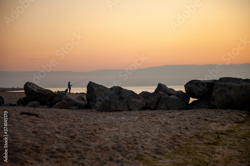 A fisherman fishing near black stones.Seascape. Waves and surf. Sunset and dawn at sea. Calm on the ocean. Beautiful sky and clouds illuminated by the sun. Natural background. Summer time.