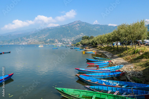 A view on Phewa Lake in Pokhara, Nepal with many colorful boats parked along it's shore. There are high Himalayan ranges in the back. Calm surface of the lake. Clear and sunny day. Serenity