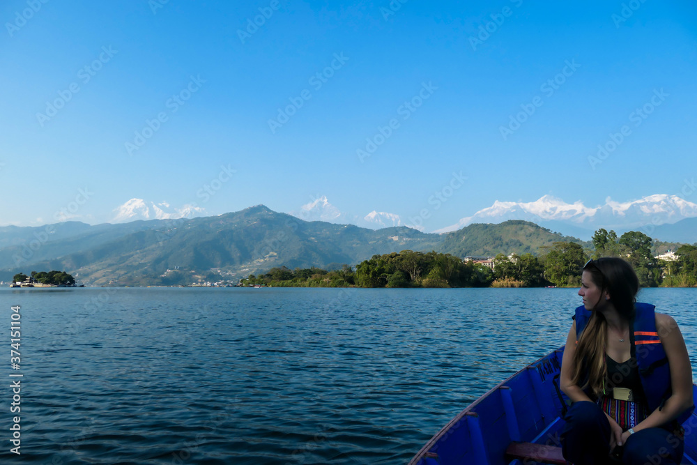 A woman sitting in a blue boat and enjoying a tour across Phewa Lake in Pokhara, Nepal. Behind her there are high, snow capped Himalayas with Mt Fishtail (Machhapuchhare) between them. Relaxation