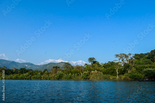 An idyllic view on Phewa Lake in Pokhara, Nepal. There are high Himalayan ranges around the lake. Calm surface of the lake. Clear and sunny day. There is a dence forest at the shore, going far inland